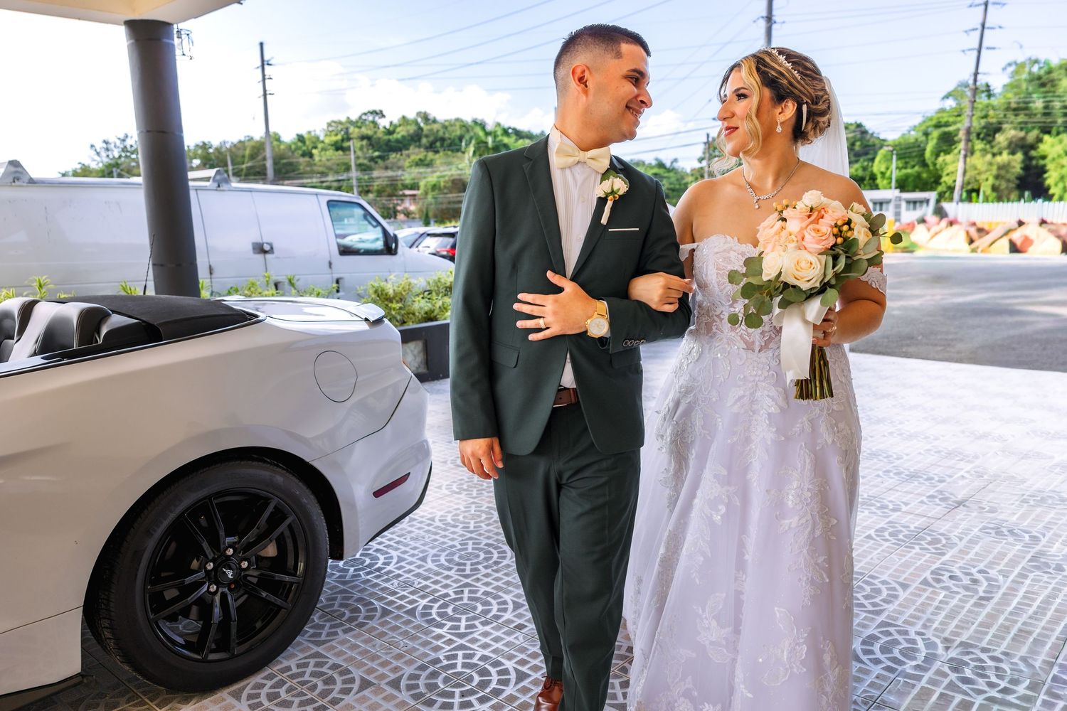 Bride and groom walking together beside a white sports car, bride holding a pink bouquet in a lace wedding gown.