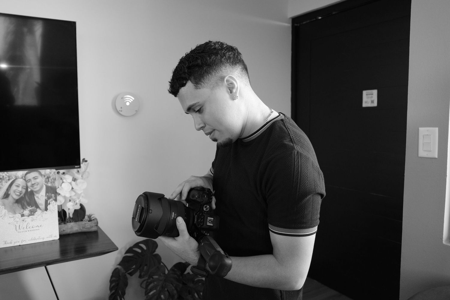 Young man in black t-shirt examines camera in monochrome indoor setting with TV and door in background.