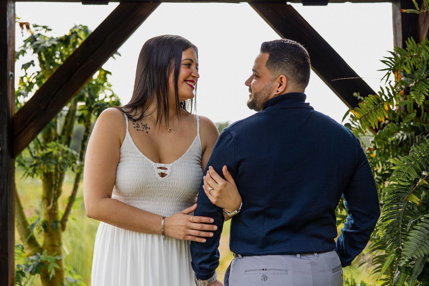 Couple sharing a romantic moment under a wooden arch surrounded by lush tropical greenery at an outdoor wedding.