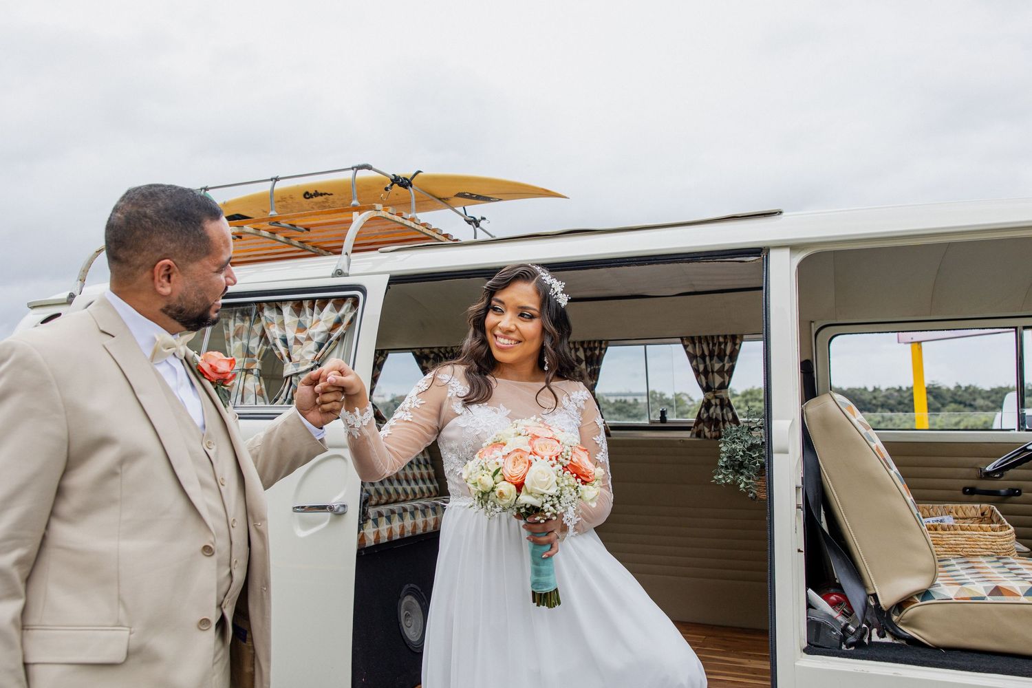Bride and groom pose by a vintage VW bus, bride holding a peach bouquet in a lace gown, groom in a beige suit.