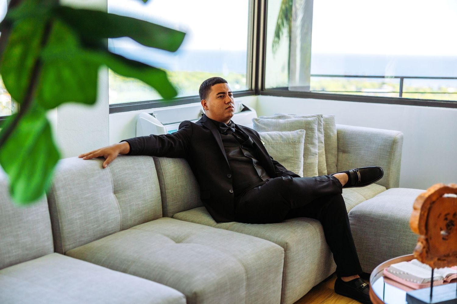 Man in black suit relaxing on gray sofa near large windows with ocean view and tropical plants nearby.