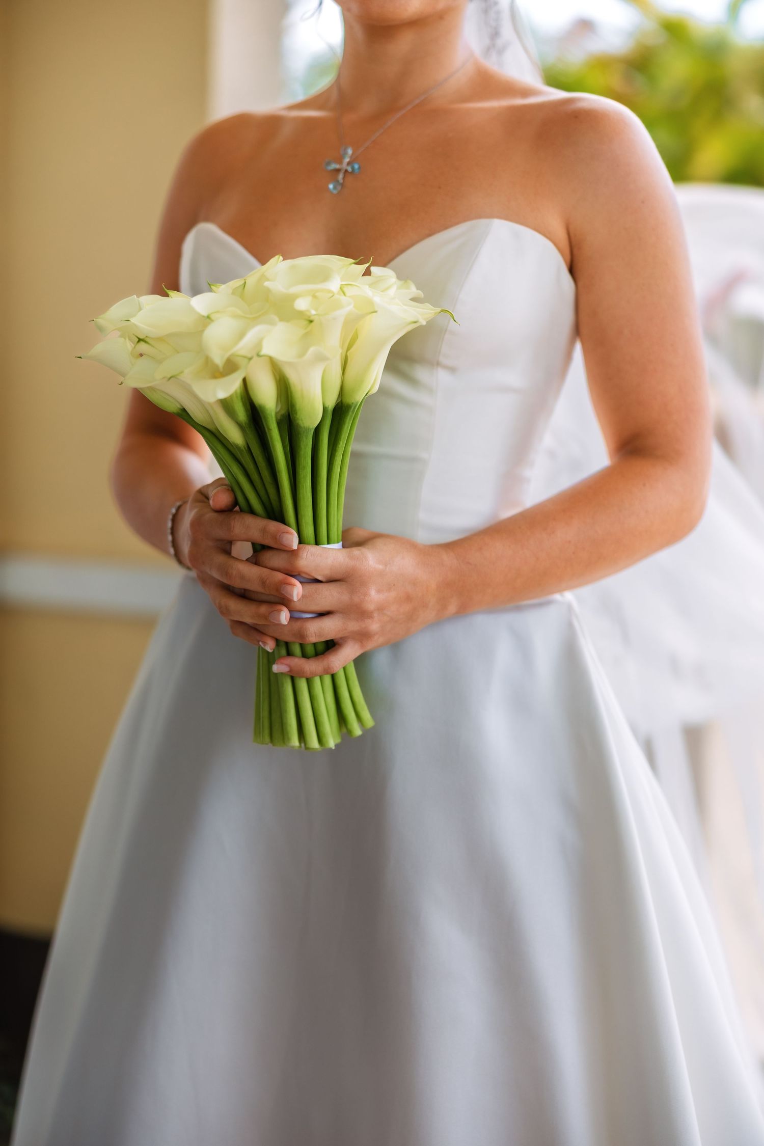 Bride in strapless white gown holding elegant white calla lily bouquet with cross necklace at wedding ceremony.