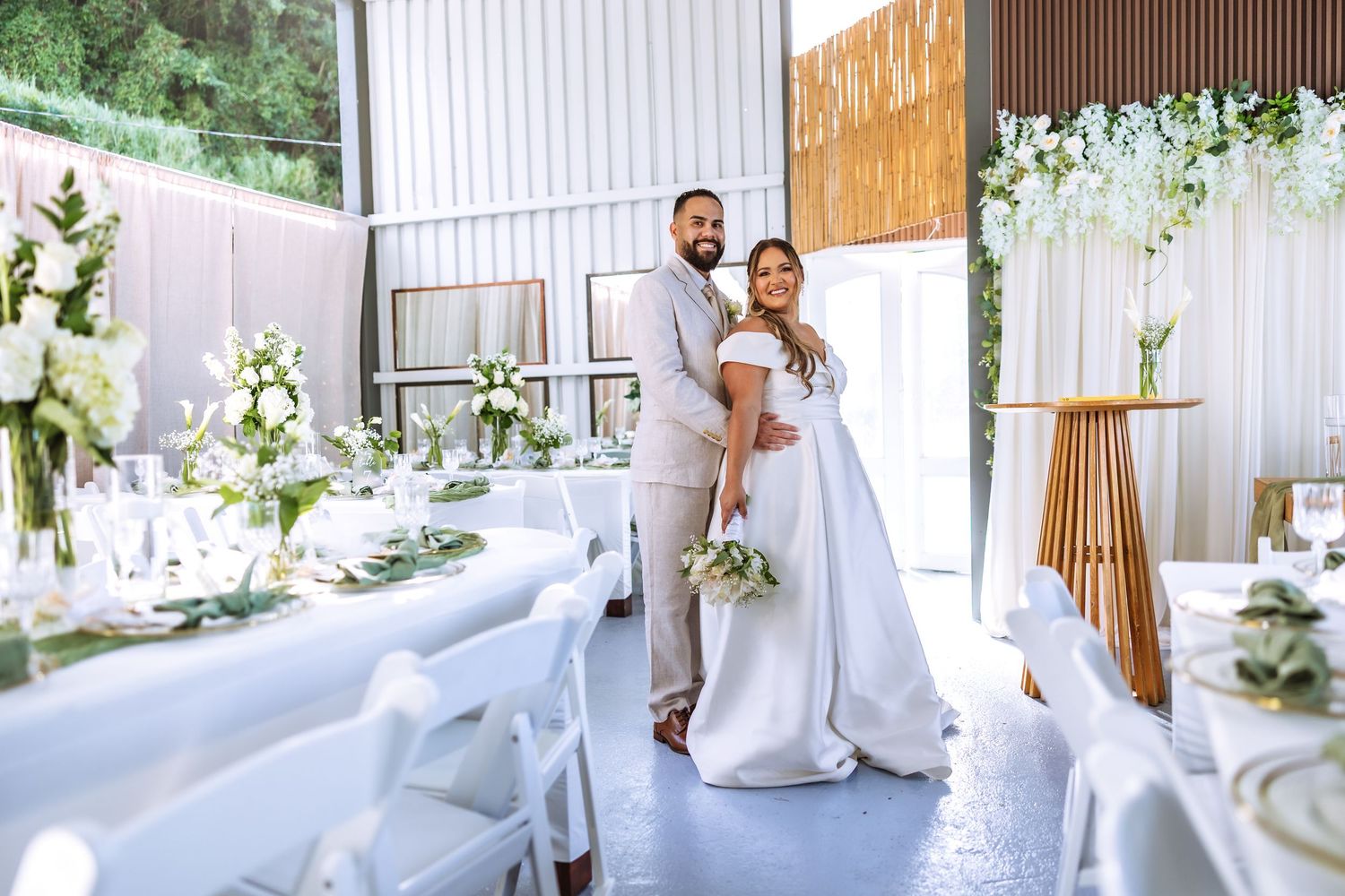 Elegant wedding reception with couple posing in white floral venue, bride in gown holding bouquet, groom in gray suit.