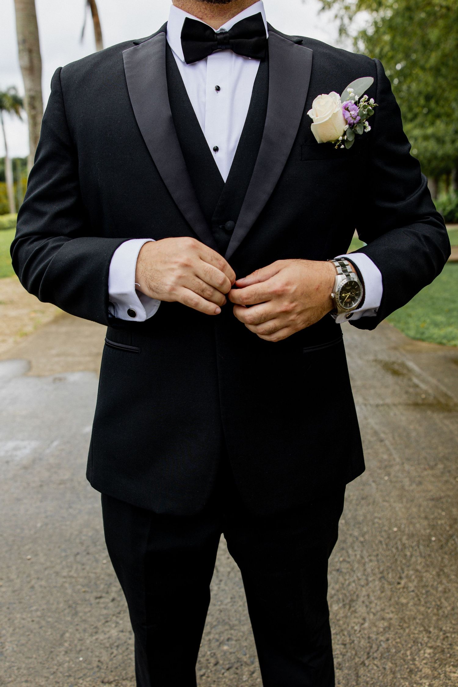Groom in black tuxedo with bow tie and white rose boutonniere, buttoning jacket before wedding ceremony.