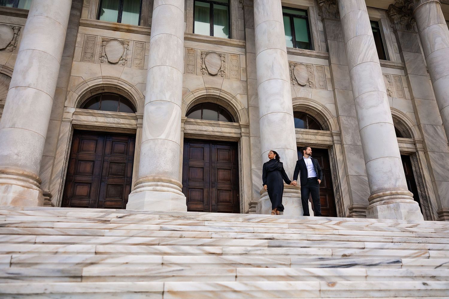 Couple holding hands descending grand marble steps of a neoclassical building with tall columns.