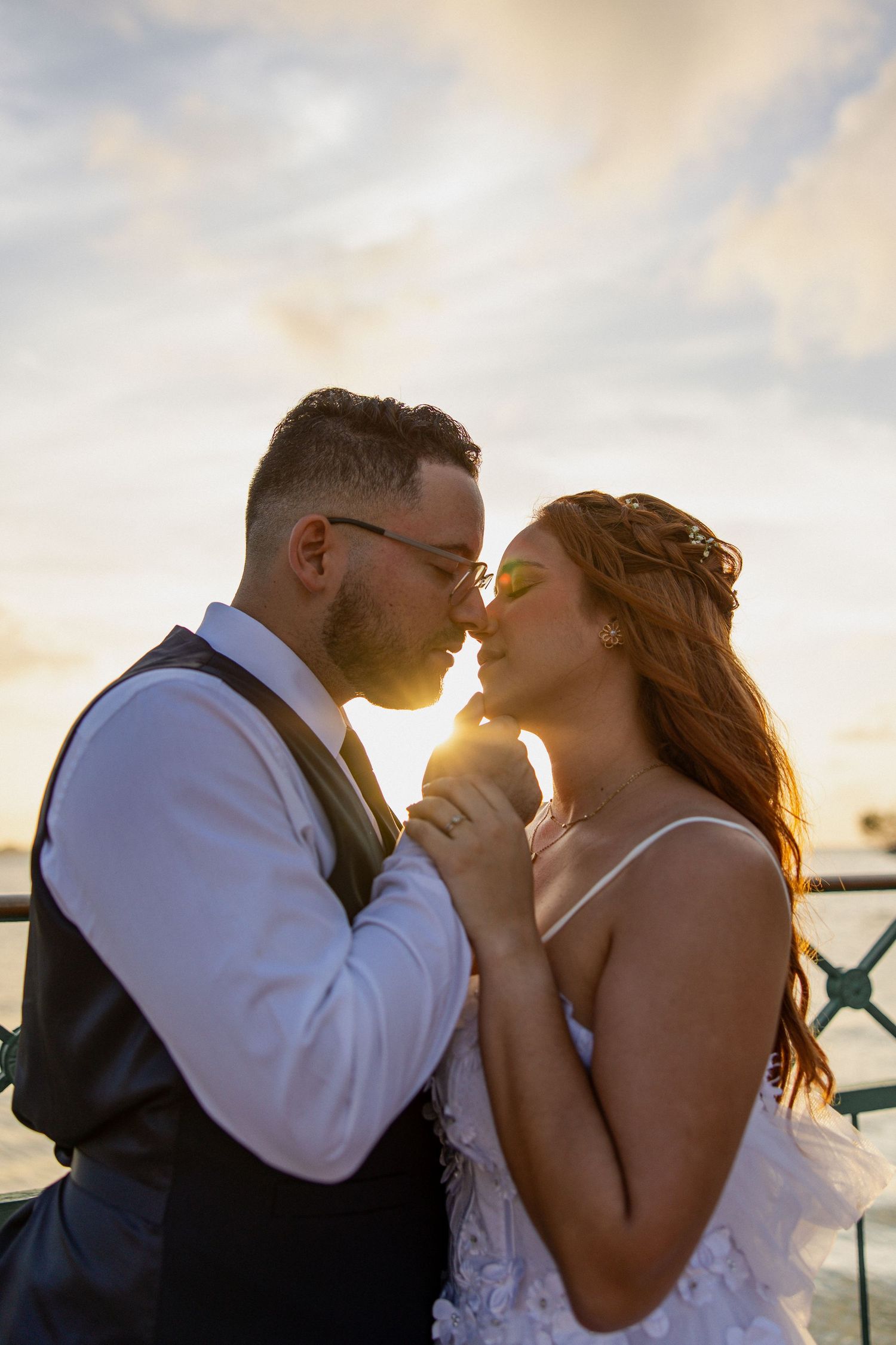 A couple shares a romantic kiss at sunset against an ocean backdrop, silhouetted by warm golden light.