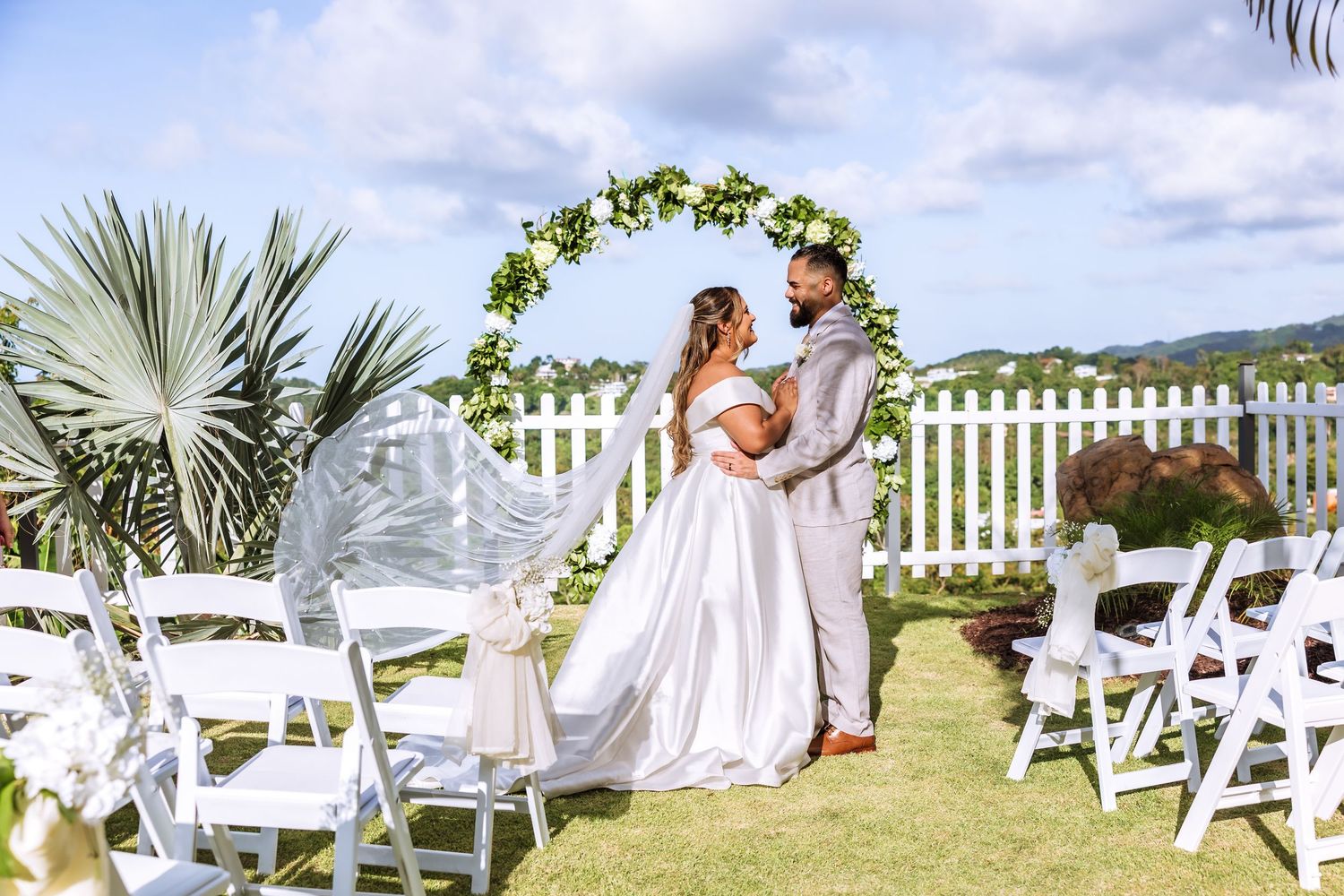 Bride and groom exchange vows under a floral arch at an outdoor wedding ceremony with white chairs.