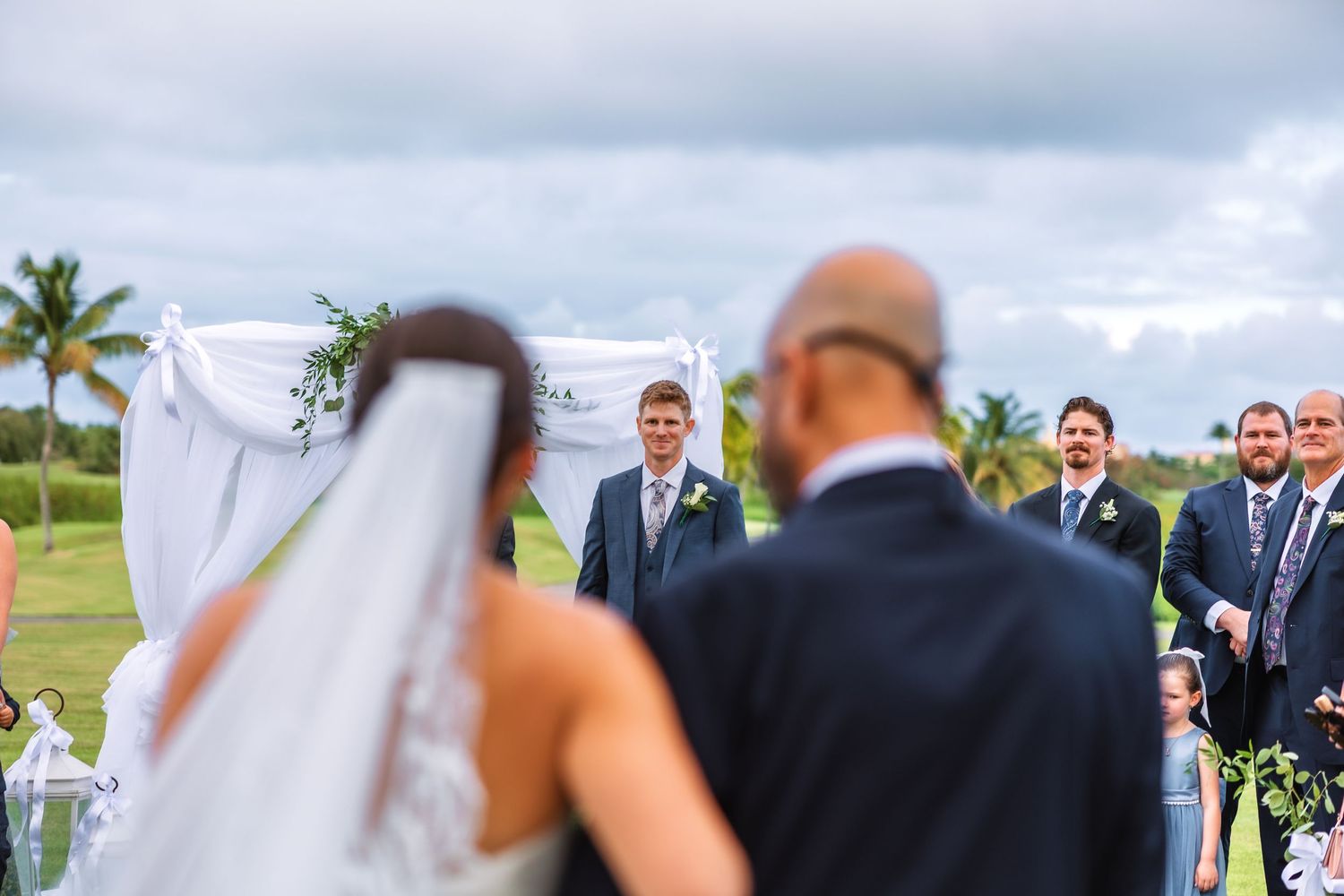 Groom smiling at bride walking down the aisle at an outdoor wedding ceremony on a golf course.