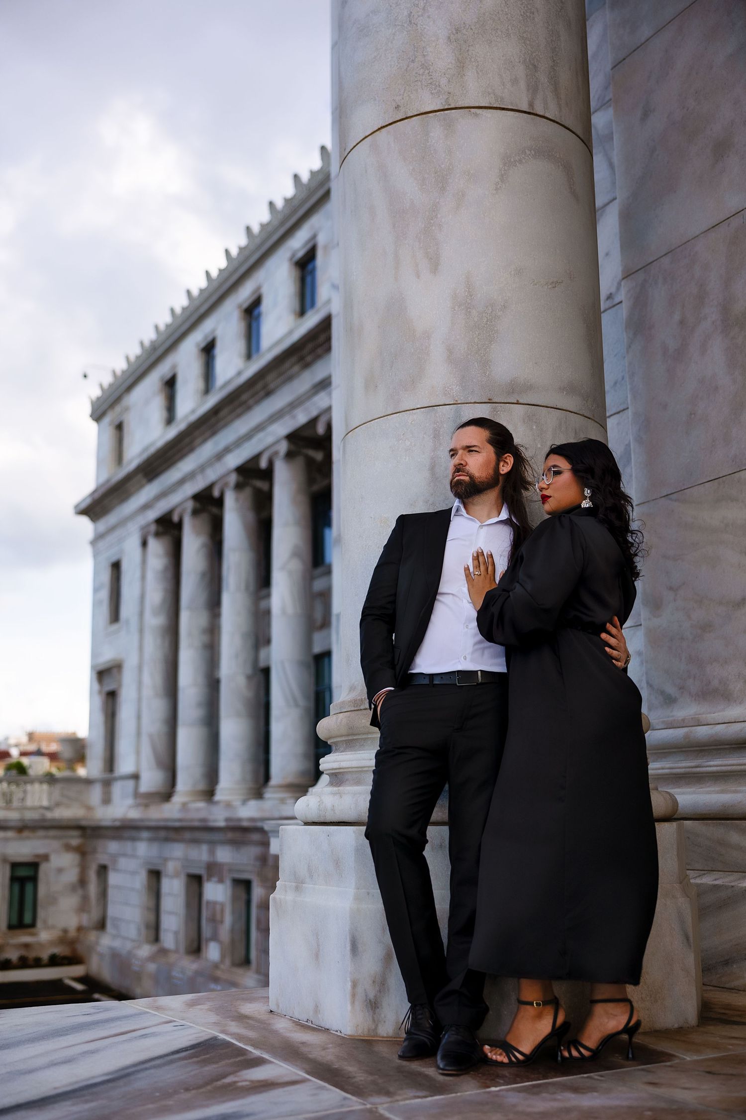 A romantic couple poses for engagement photos between classic white columns of a historic building.