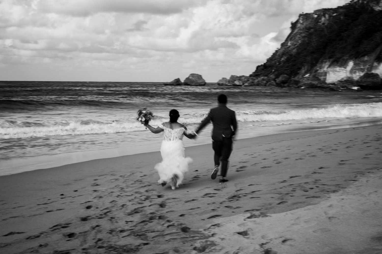 Black and white panoramic sequence showing people playing with a dog on a beach with waves and cliffs in the background.