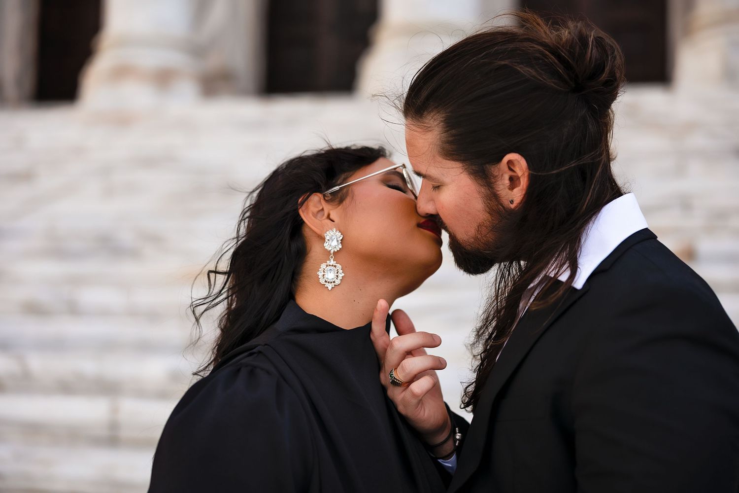 Romantic couple sharing a kiss outdoors, woman wearing elegant earrings and black outfit, man with long hair in bun.
