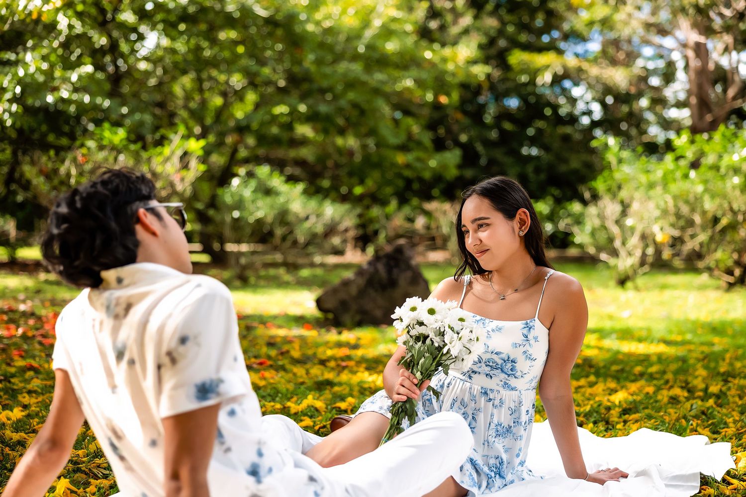 Two people in floral outfits sit on grass in a sunny park, woman holding white flowers facing her companion.