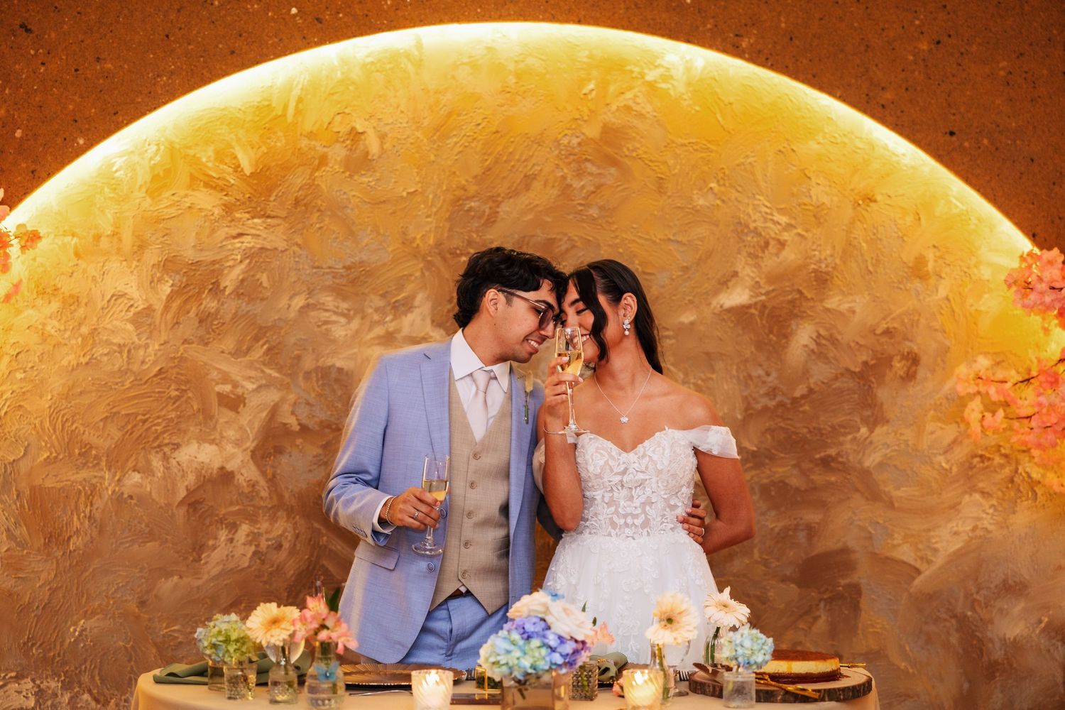 Bride and groom toasting champagne at sweetheart table with golden moon backdrop and floral centerpieces.