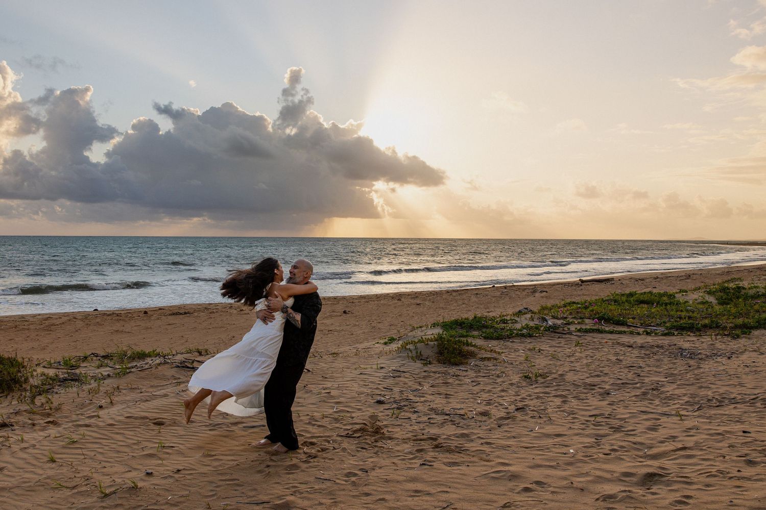 Couple dancing on a sandy beach at sunset with dramatic rays of light breaking through clouds over the ocean.