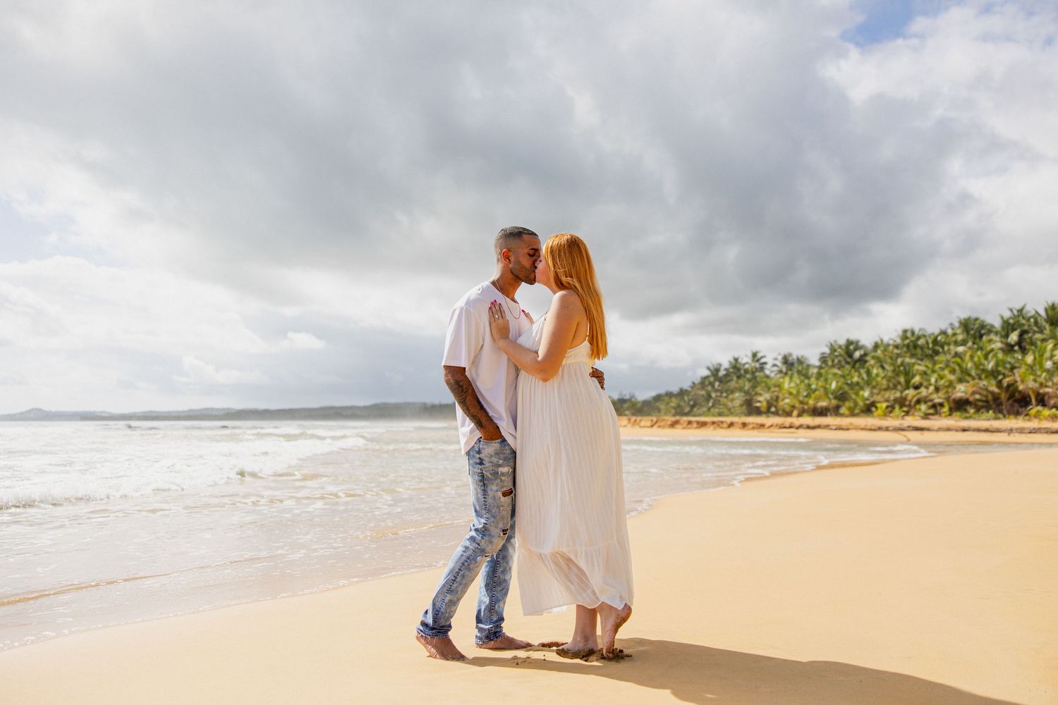 Romantic couple embracing on a tropical beach with palm trees, ocean waves, and cloudy sky in the background.