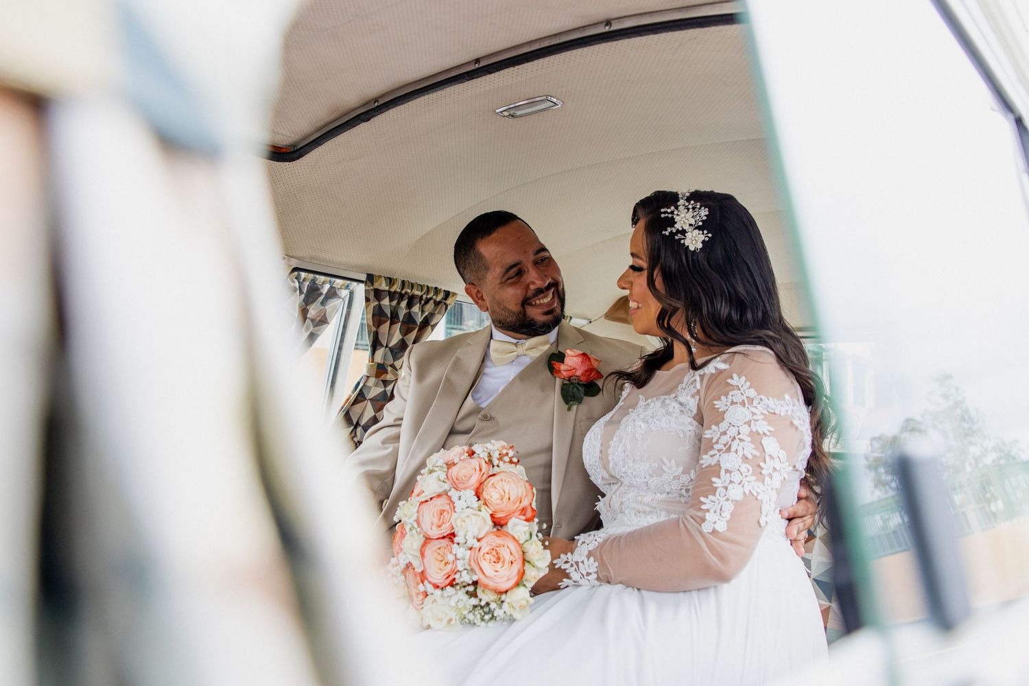 Bride and groom share a romantic moment inside a vintage car, bride holding a peach rose bouquet in wedding attire.