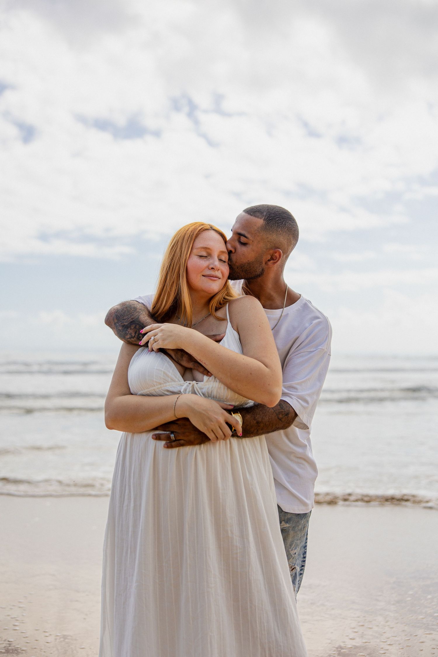 A couple embraces on a beach, man kissing woman's cheek as she holds a small dog, ocean waves in background.