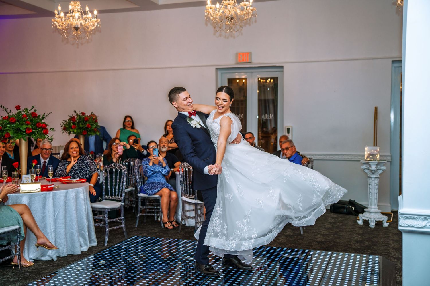 Bride and groom share their first dance at elegant wedding reception with chandeliers and guests seated nearby.