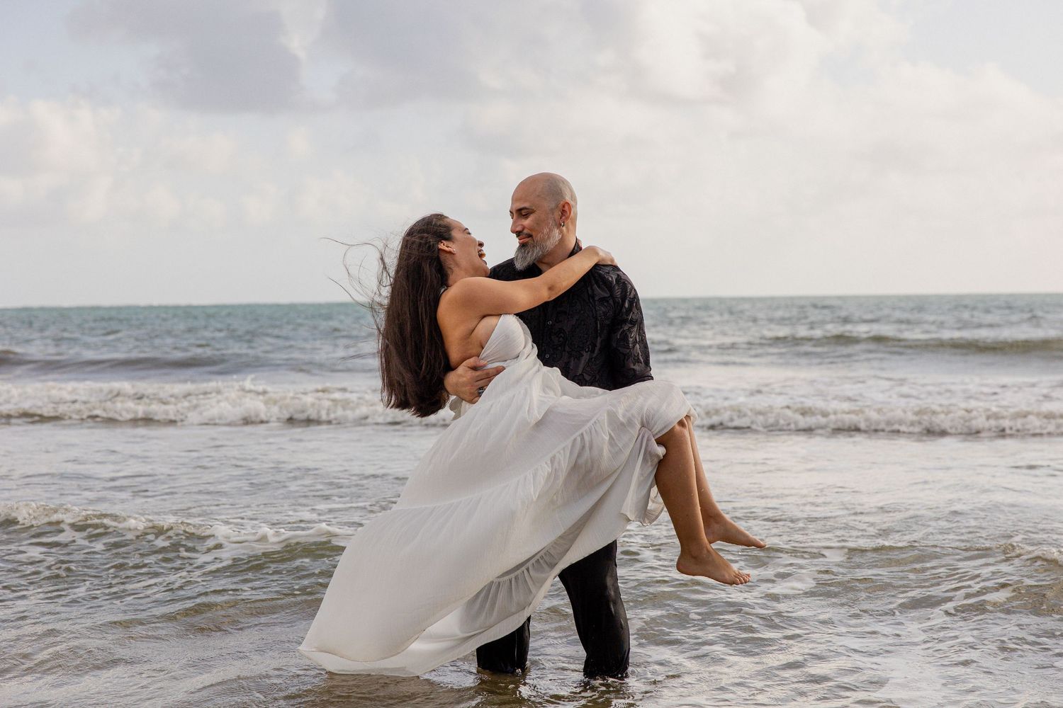 Couple in formal wedding attire embracing on a beach with waves crashing behind them at sunset.
