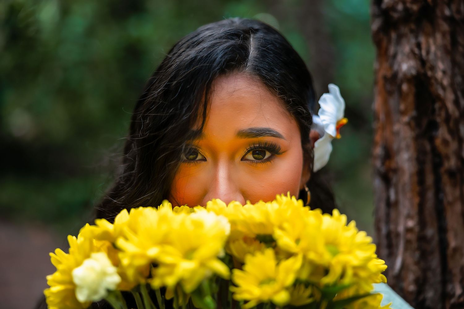 Young woman with dark hair and a white flower accessory holds a bouquet of bright yellow chrysanthemums near her face outdoors.