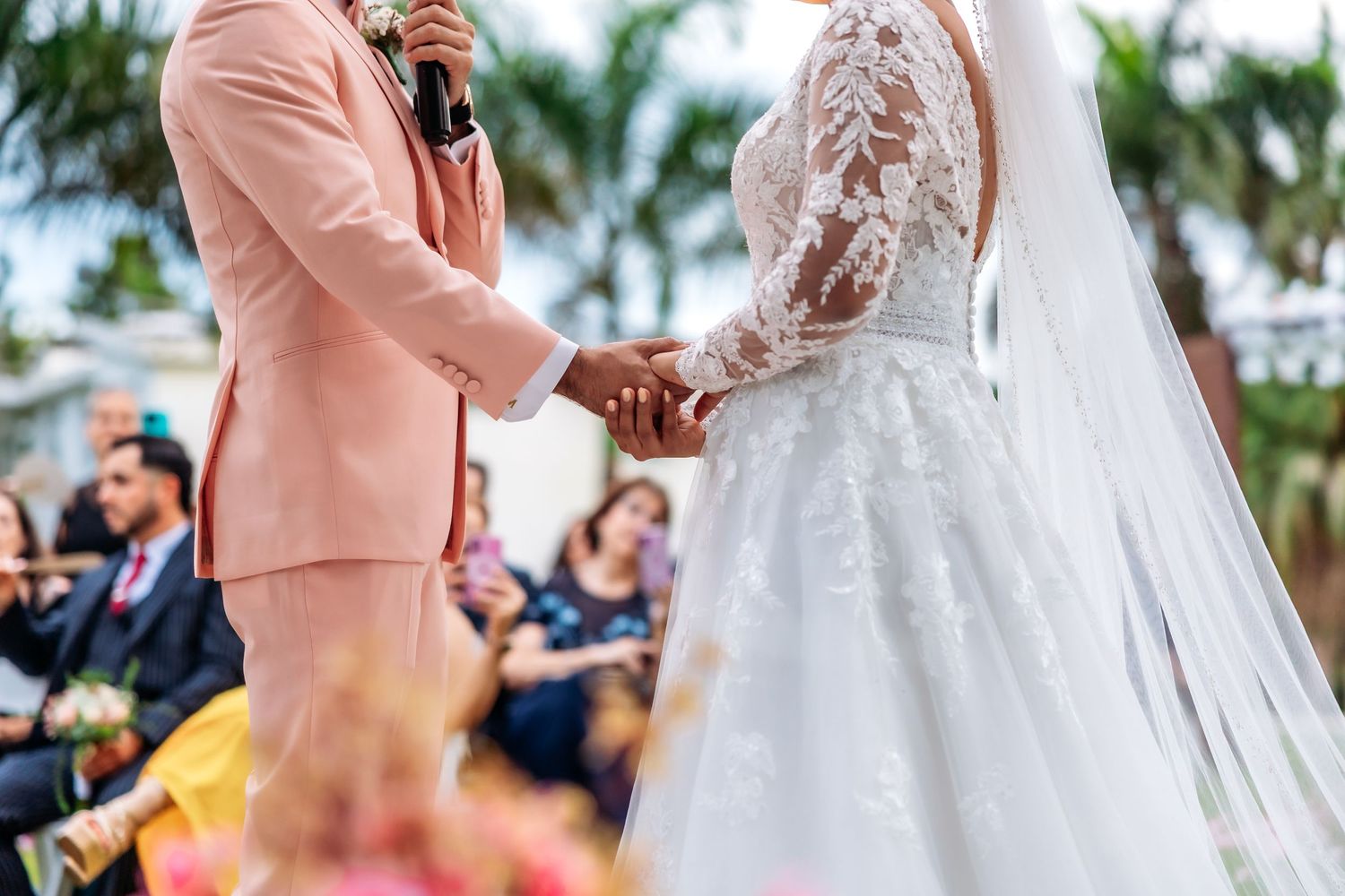 Bride and groom holding hands at outdoor wedding ceremony, bride in lace gown, groom in pink suit.