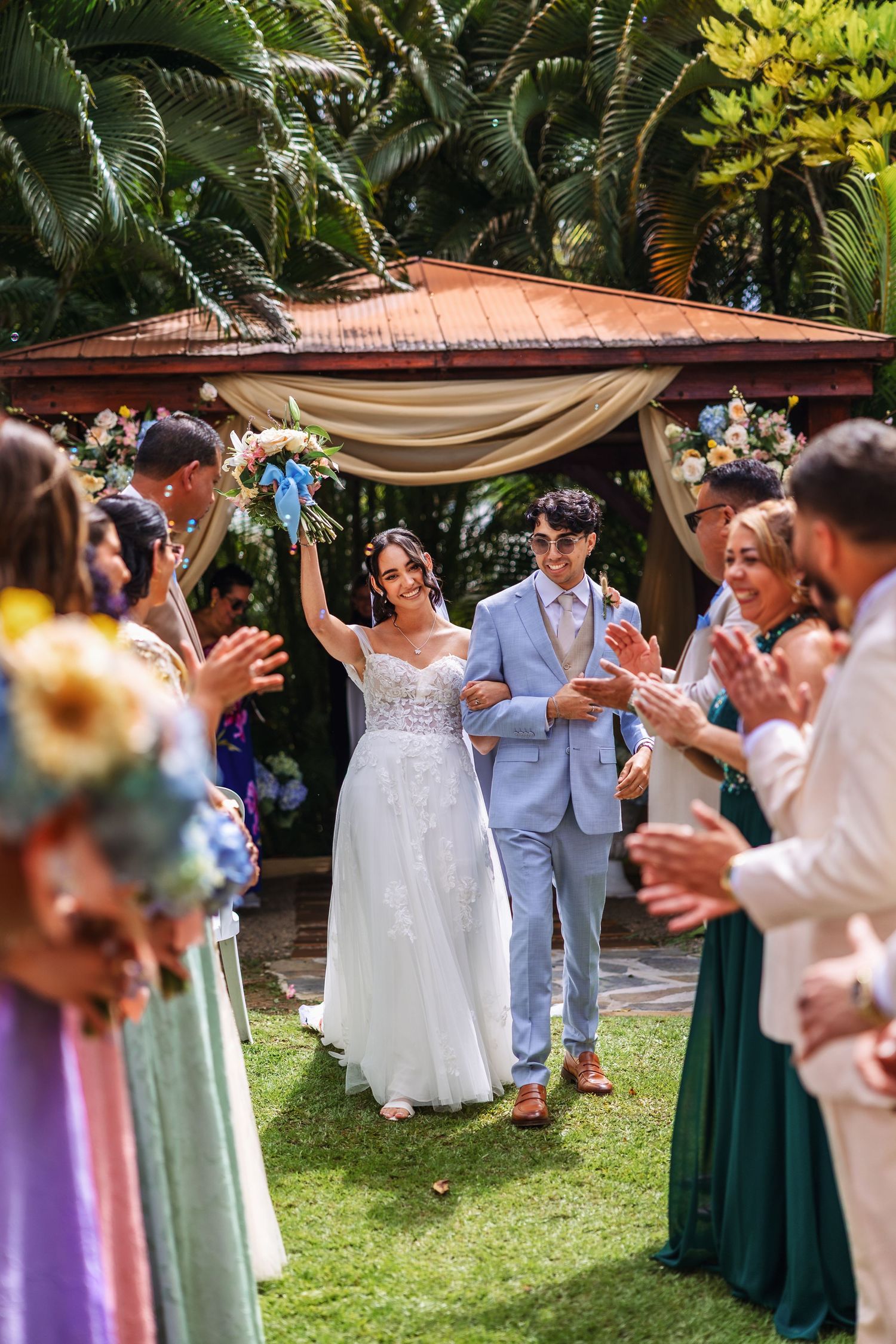 Bride and groom walk down outdoor aisle, guests clapping, lush tropical greenery and floral arch backdrop.