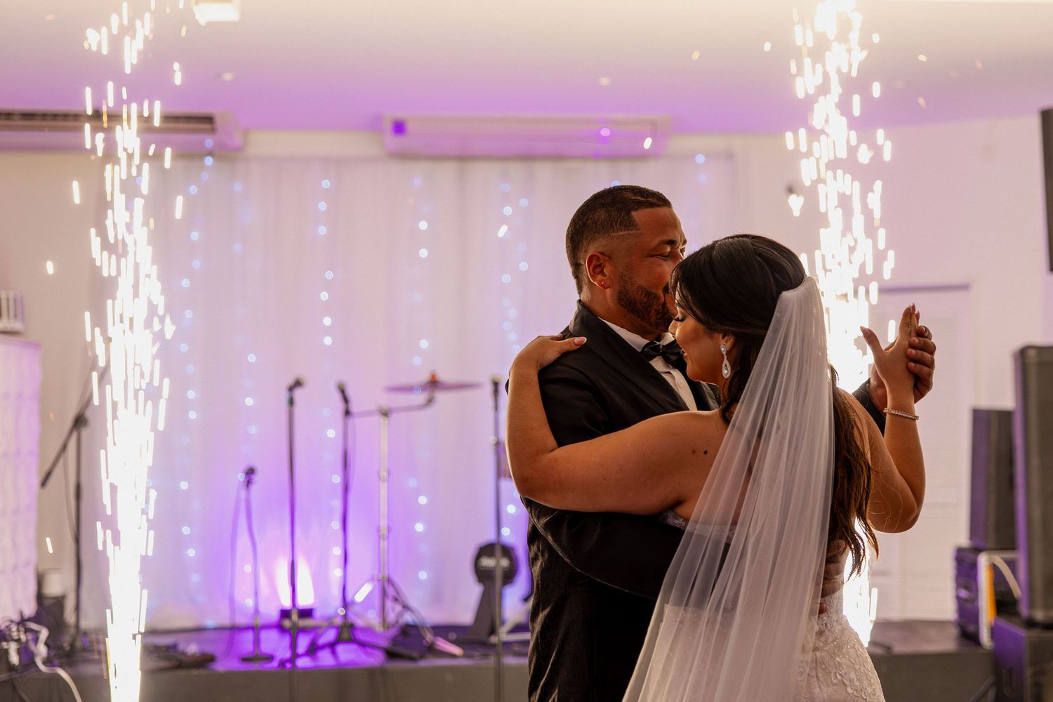 Sparklers illuminate a wedding reception dance floor as a couple shares their first dance in a white decorated ballroom.
