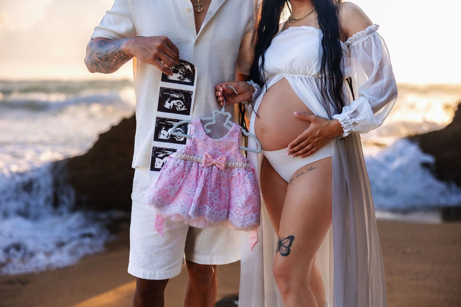 Pregnant couple on beach holding ultrasound photos and pink baby dress during golden hour maternity photoshoot.