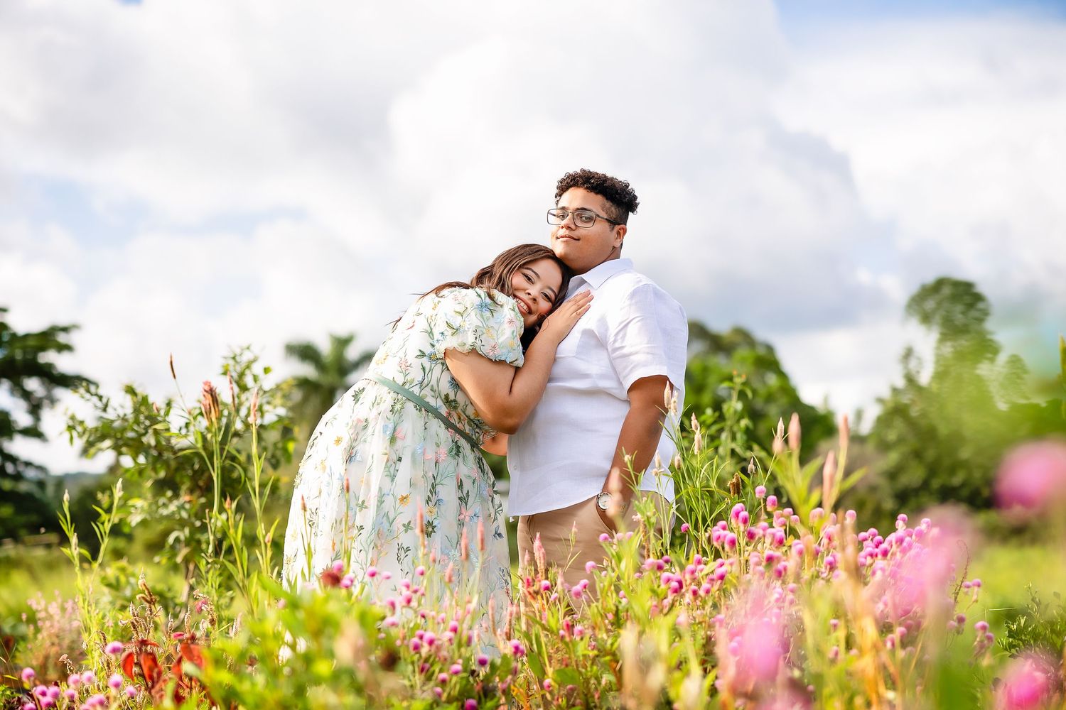 Couple embracing in a vibrant flower field under a cloudy sky, woman in floral dress leaning on man in white shirt.