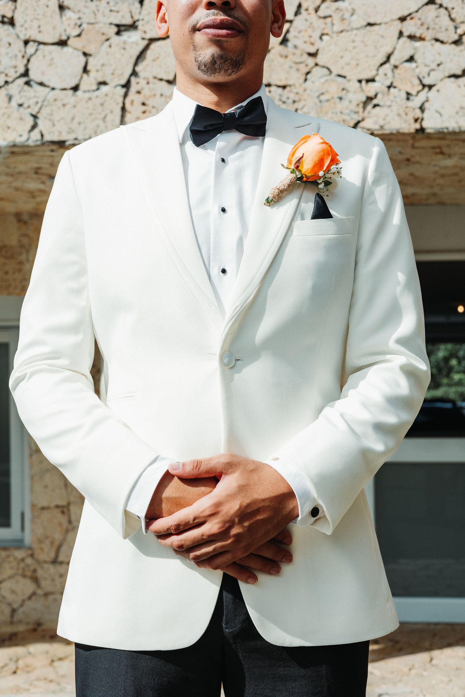 Close-up details of a white tuxedo jacket with orange boutonniere against a stone wall background.