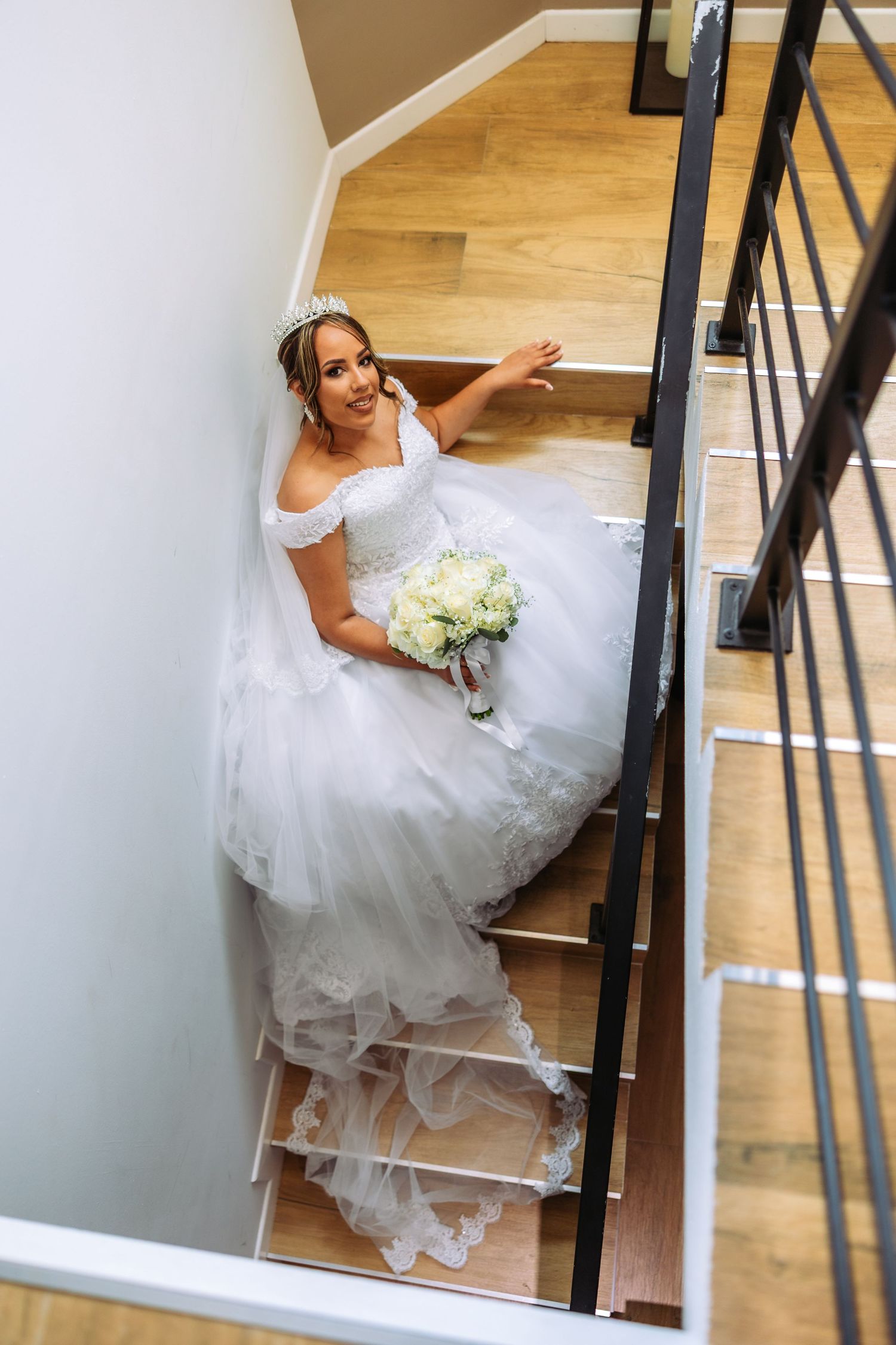 Bride in white ball gown with tiara holds bouquet on wooden staircase, looking up with a radiant smile.