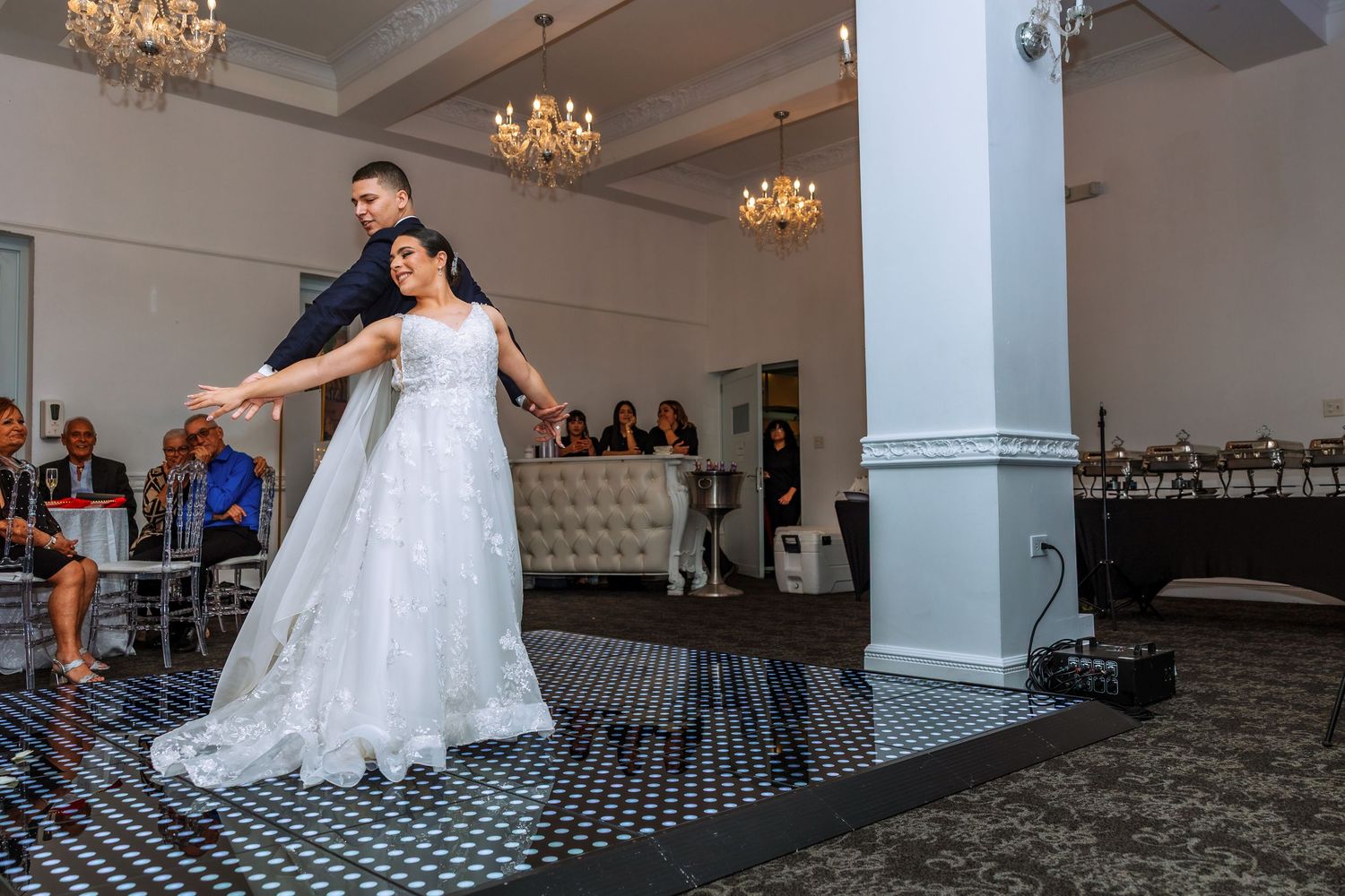 A couple performs their first dance on a checkered dance floor in an elegant ballroom with crystal chandeliers.
