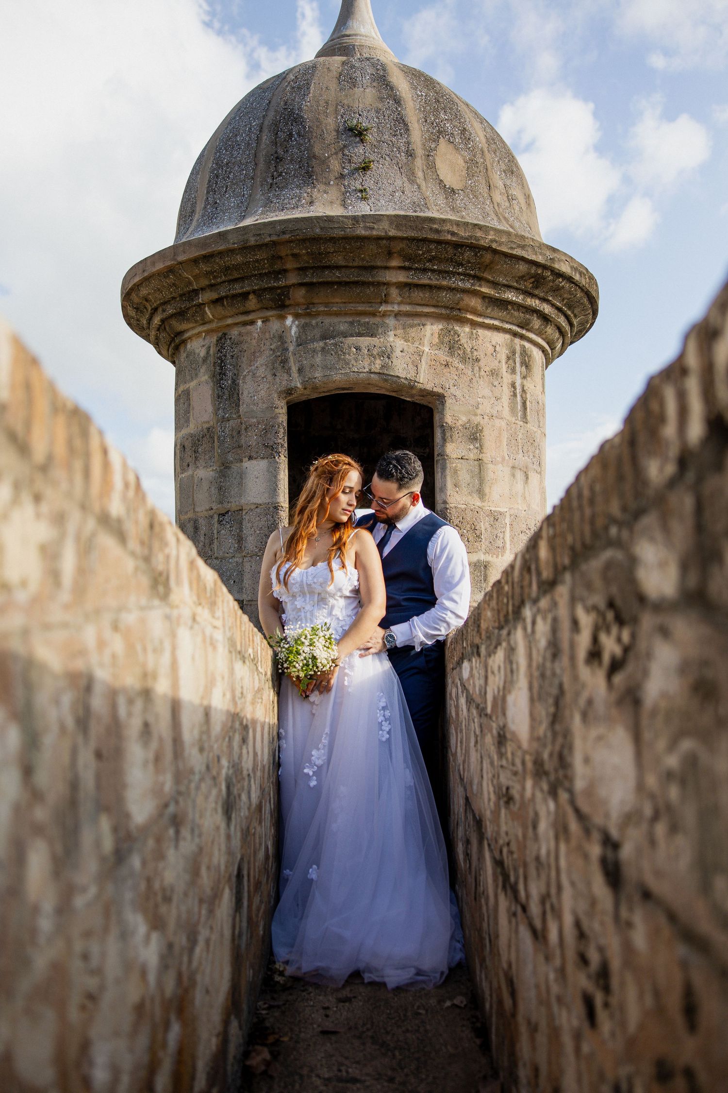 Bride and groom embrace in a historic stone fort turret, bride holding a bouquet in a flowing white gown.