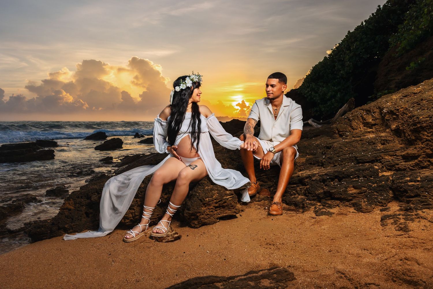 Couple in white outfits posing on rocky beach at sunset with ocean waves and golden sky backdrop.