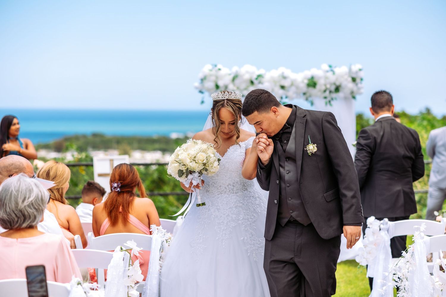 Groom kissing bride's hand at outdoor wedding ceremony with ocean view and floral arch backdrop.