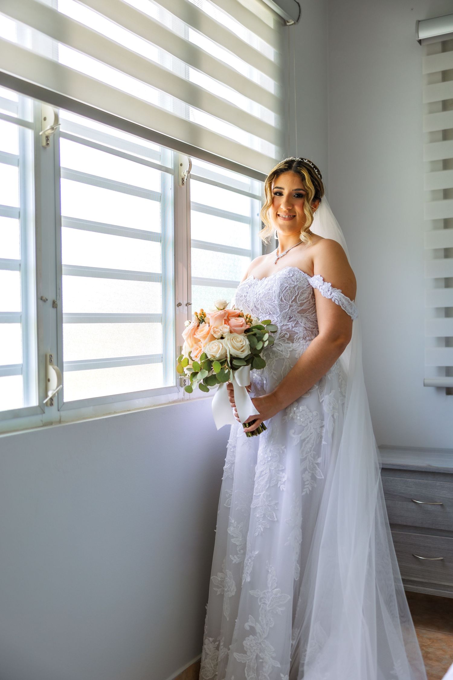 Bride in white lace off-shoulder gown holding peach and white bouquet, standing by window with natural light.
