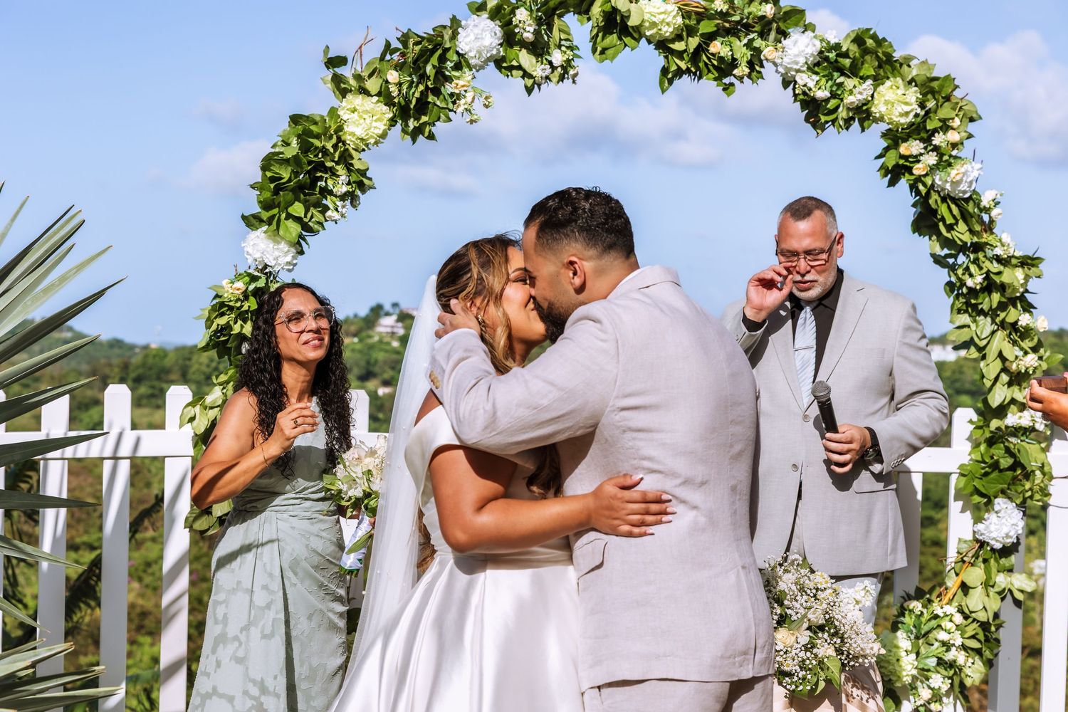 Newlyweds share first kiss under floral arch at outdoor wedding ceremony with guests celebrating nearby.