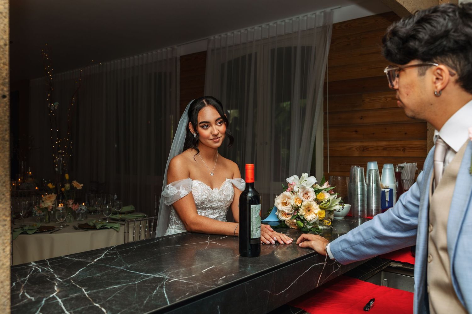 Bride and groom at marble bar with wine bottle and floral bouquet during elegant wedding reception.