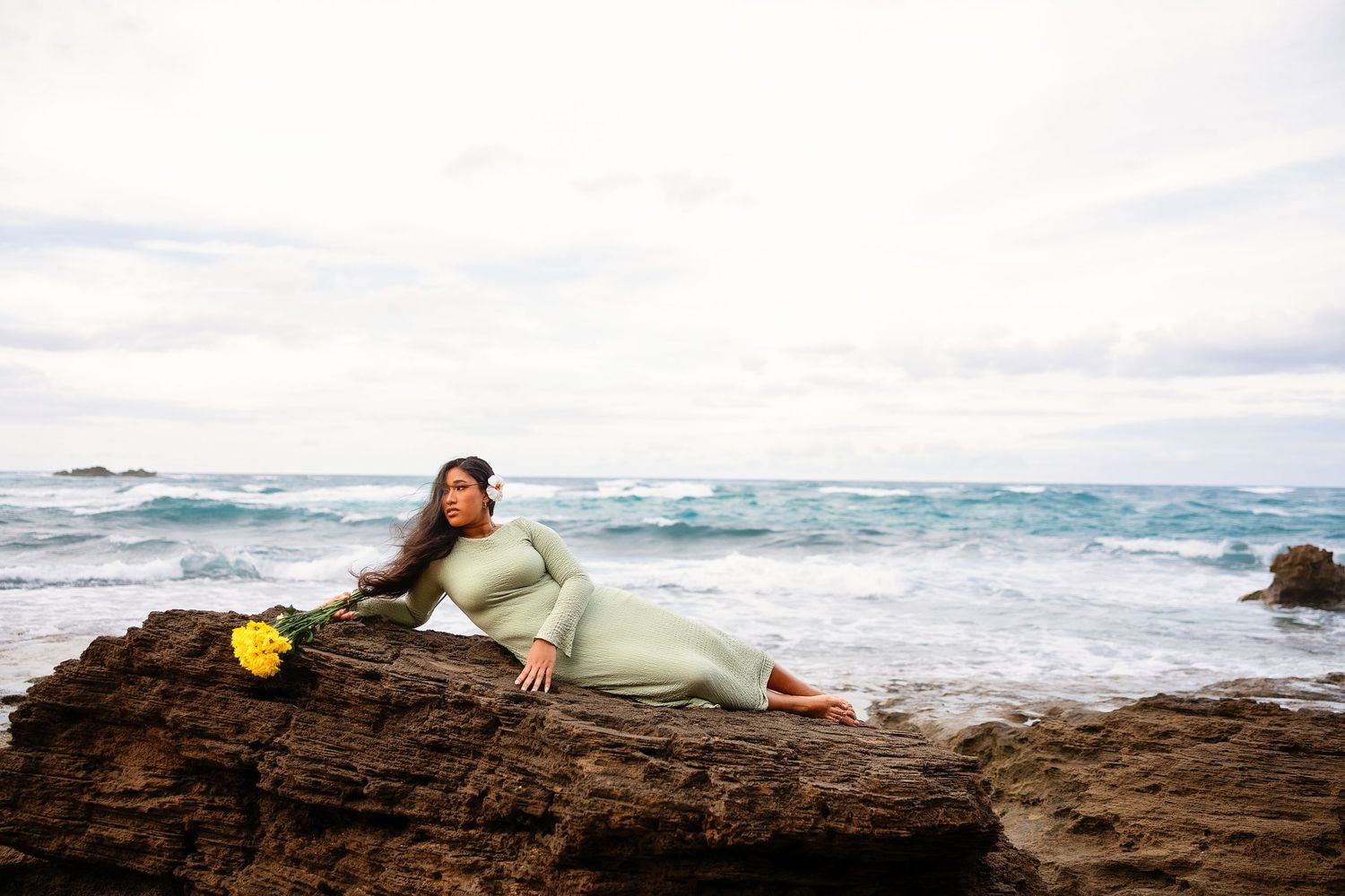 Pregnant woman in sage green dress reclining on coastal rocks with yellow flowers, ocean waves in background.