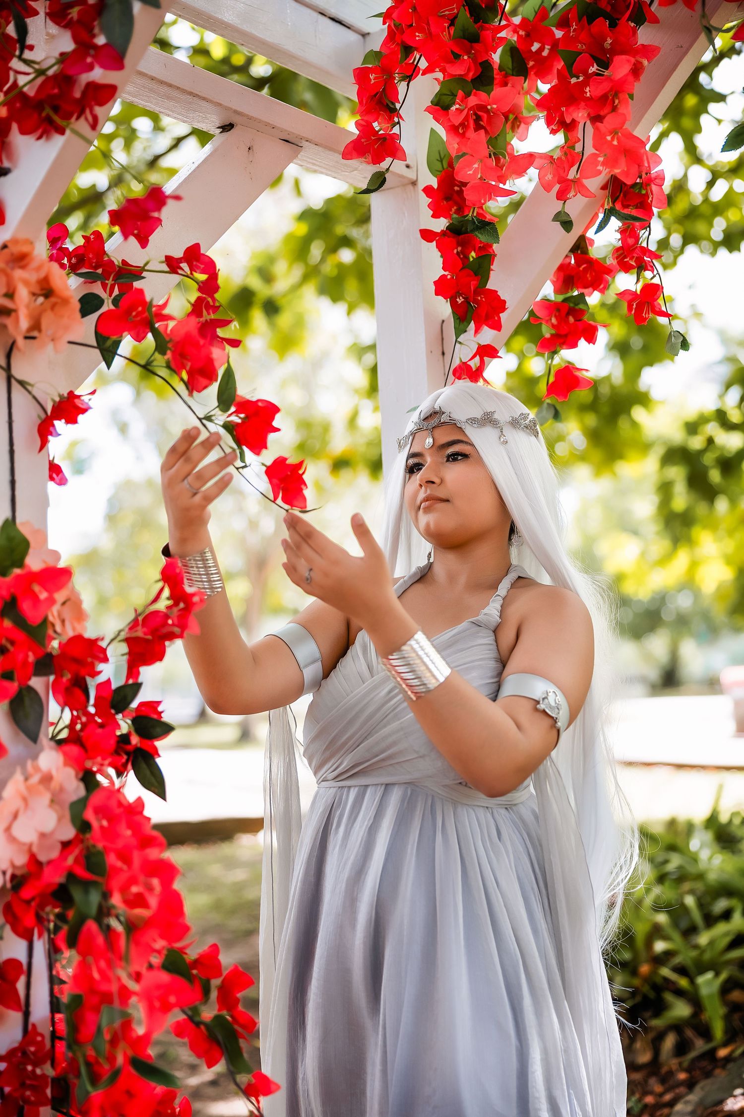 Woman in silver dress and white wig poses gracefully among vibrant red bougainvillea flowers under a white garden arbor.