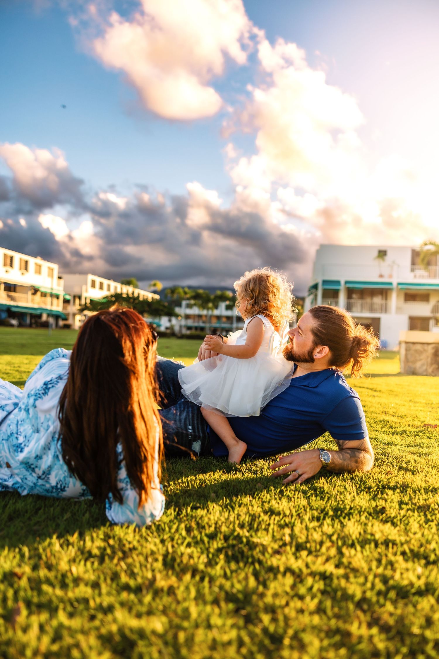 Family relaxing on green grass at sunset, mother with red hair, bearded father, and curly-haired toddler in white dress.
