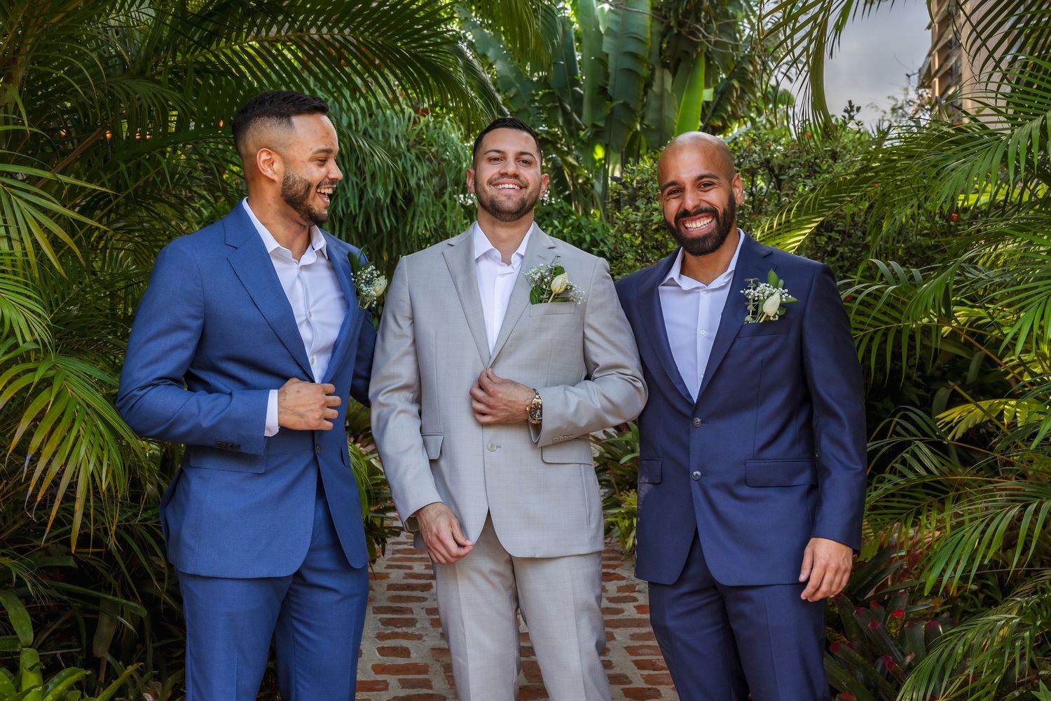 Three groomsmen in blue and gray suits with boutonnieres smiling outdoors among lush tropical greenery.