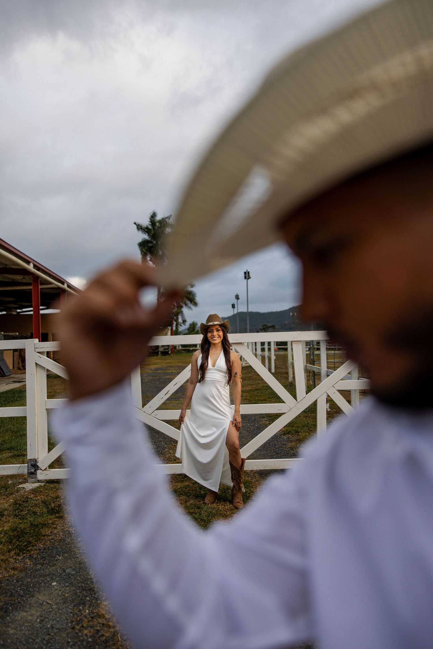 Man in white shirt tipping cowboy hat with woman in white dress standing by white farm fence in background.