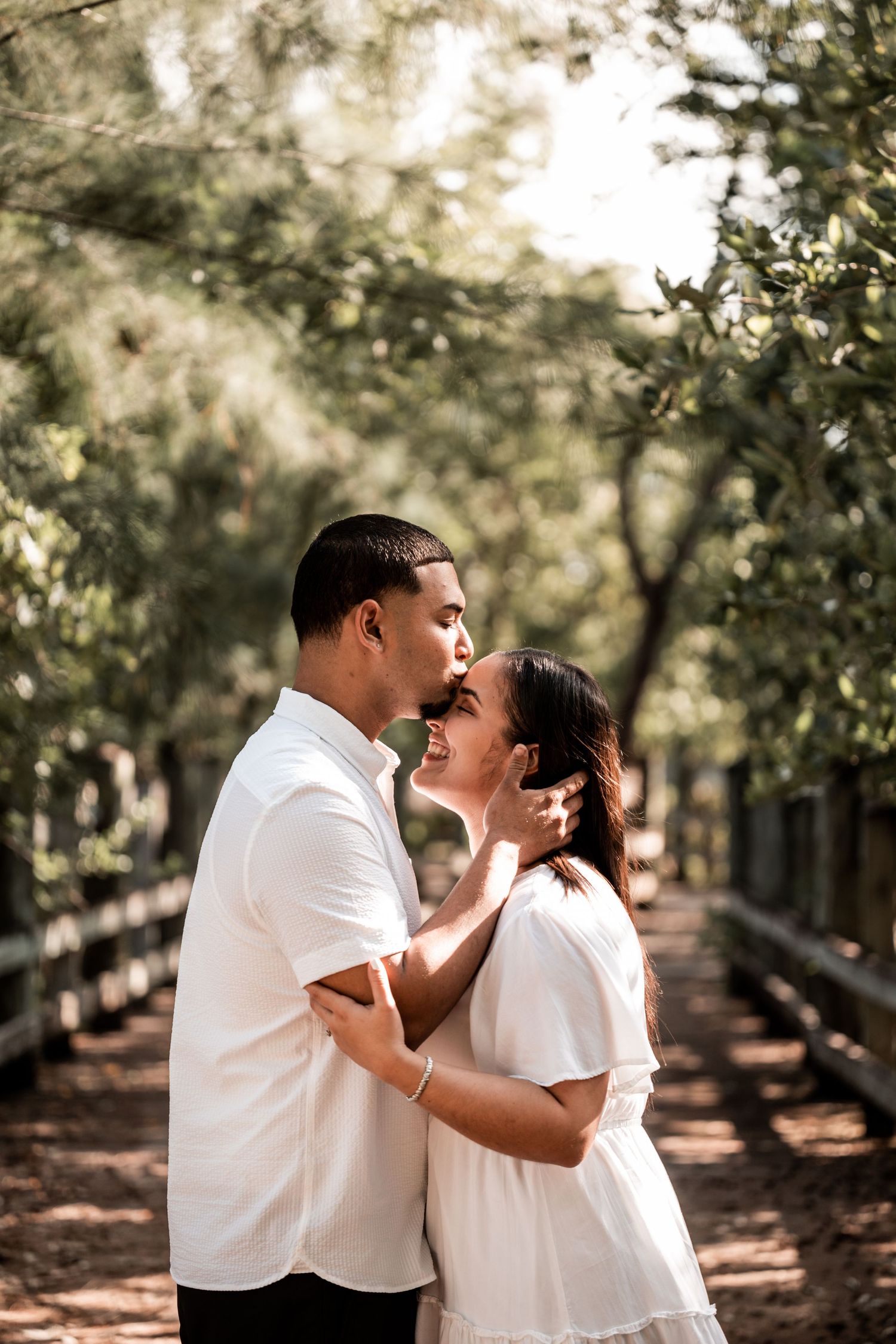 Couple in white outfits sharing a romantic kiss on a tree-lined path in a lush green park.