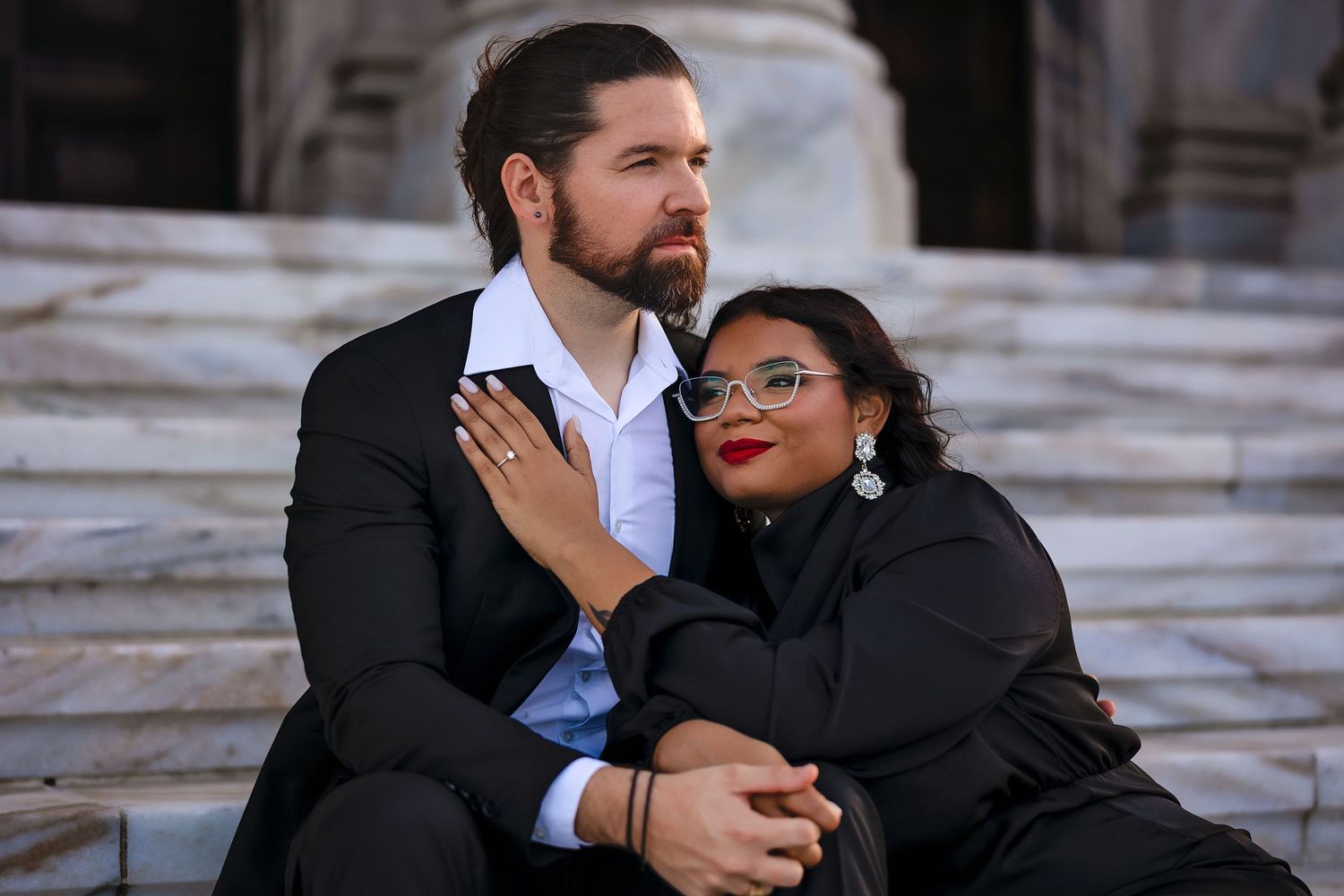 Couple in black formal wear sitting on stone steps, woman leaning on man's shoulder, engagement ring visible.