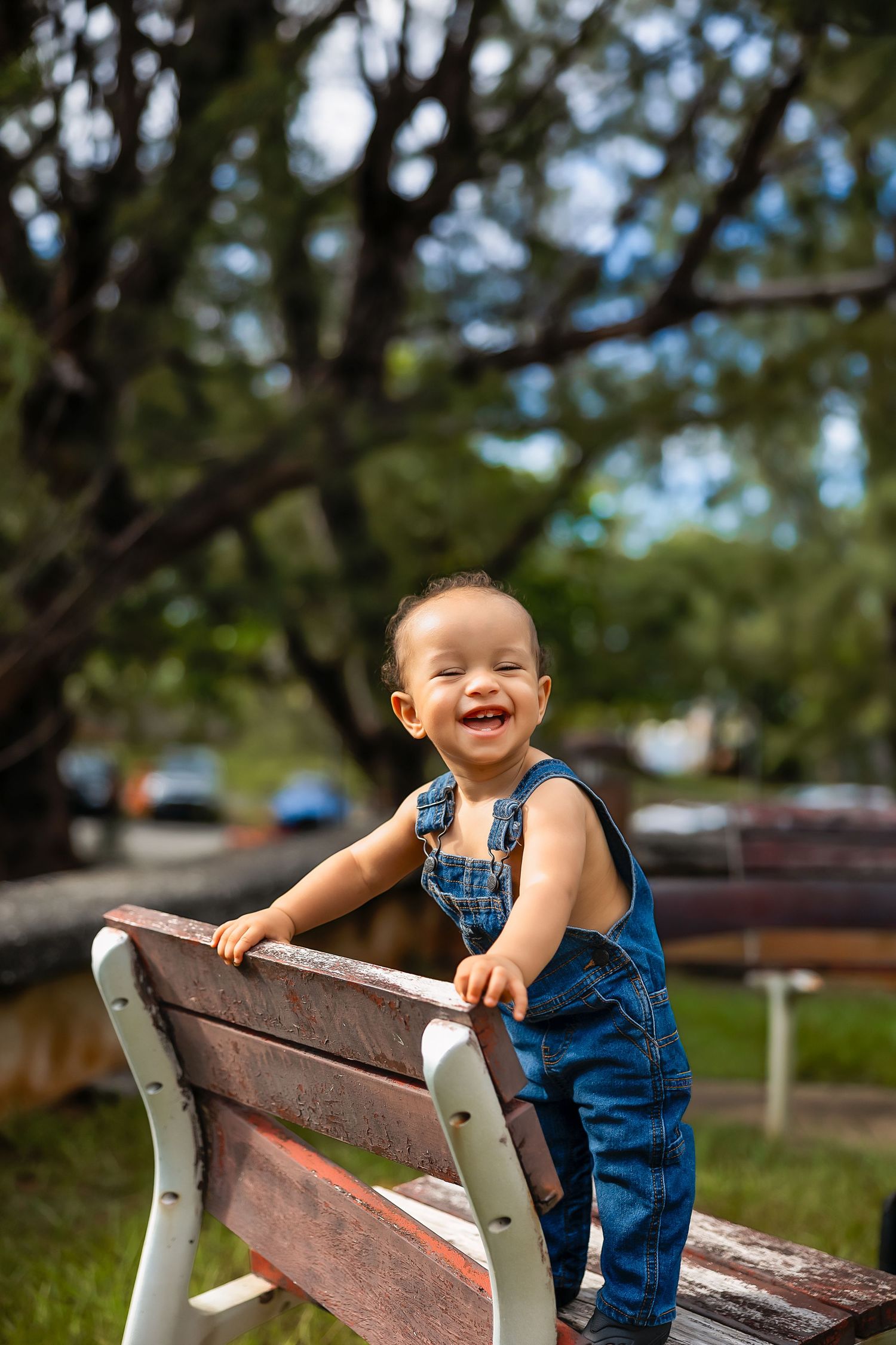Smiling toddler in denim overalls standing on a wooden park bench outdoors with green trees in the background.