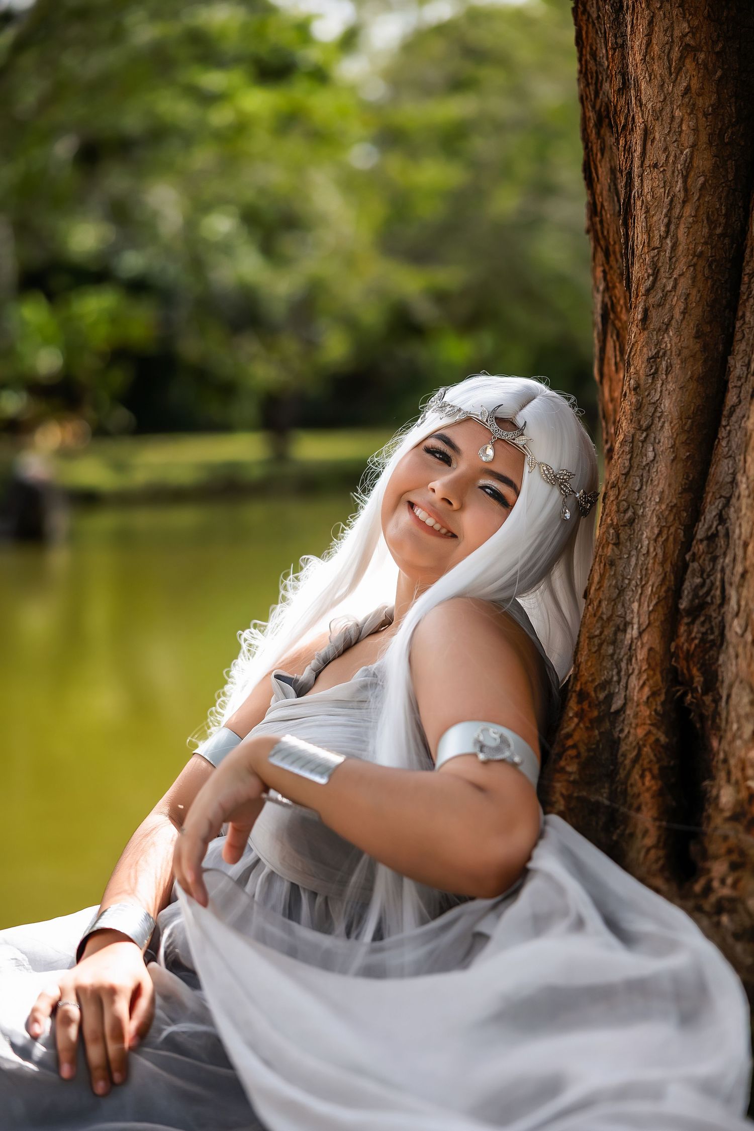 Woman in white fantasy elf costume with silver hair sits against a tree in a lush green park setting.