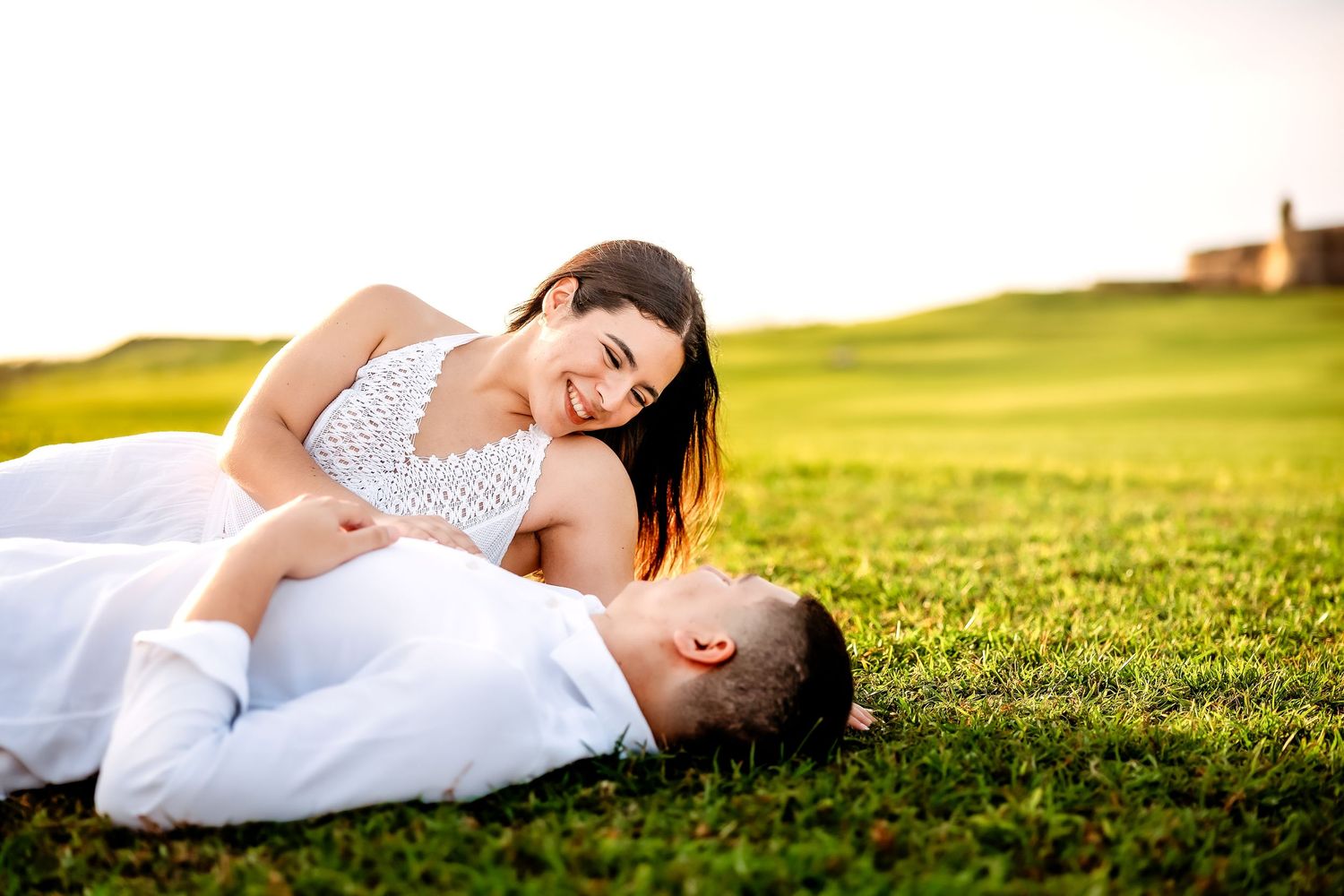 A couple in white clothing lies together on lush green grass during a sunny outdoor photoshoot.