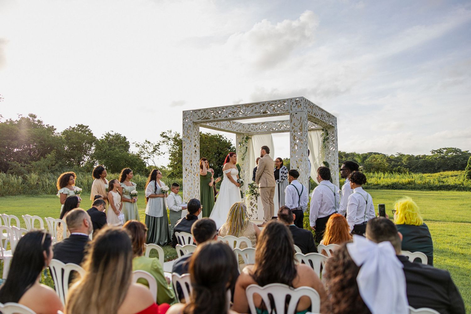 Guests gather outdoors for a rustic countryside wedding ceremony under a white wooden arch surrounded by greenery.