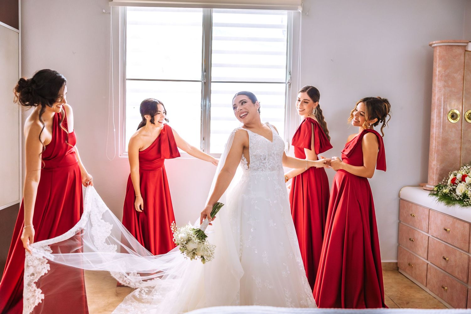 A bride in a white wedding gown stands with bridesmaids wearing matching red dresses in a sunlit room.